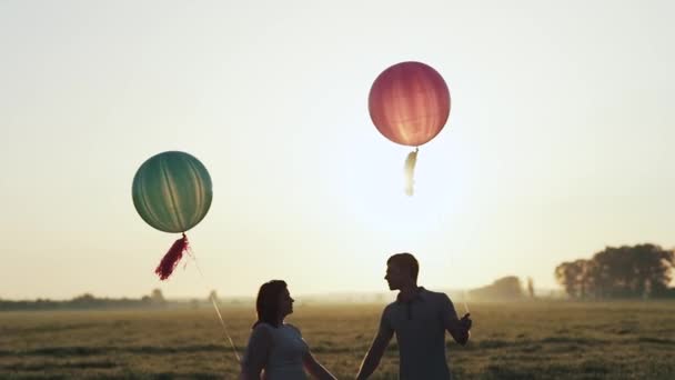 Пара с гелиевыми шариками, идущими в поле и целующимися на закате — стоковое видео