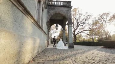 Düğün çifti tarihi bir sarayla parkta yürüyor. Gelinlik. Yeşil ağaçlar ve arka planda bir bina.