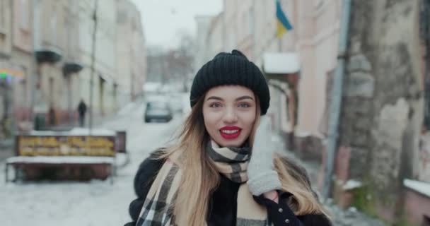 Attraktiv tjej i en svart päls med röda läppar går nerför gatan i en stad, än vänder sig till kamera och leenden. — Stockvideo