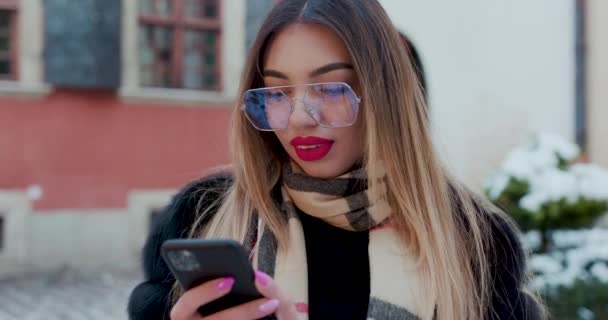 Woman with glasses look at camera poses in city center Beautiful hair attractive smile girl. Close up shot on hands using phone during winter time. Technology, winter and holiday concept. — Stockvideo