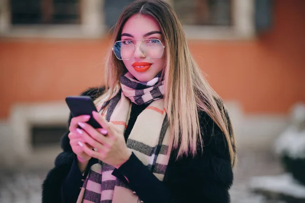Portret kobiety w okularach pozuje w centrum miasta Piękne włosy atrakcyjny uśmiech dziewczyny. Blisko strzał na ręce przy użyciu telefonu w okresie zimowym. Koncepcja technologii, zimy i wakacji. — Zdjęcie stockowe