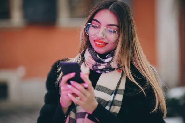 Portret młodej kobiety w okularach pozuje na Smart Phone w centrum miasta Piękne włosy atrakcyjny uśmiech dziewczyny. Blisko strzał na ręce przy użyciu telefonu w okresie zimowym. Koncepcja zimy i wakacji — Zdjęcie stockowe