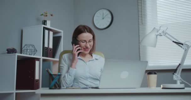 Mujer casual joven sentada en el escritorio usando el ordenador portátil, respondiendo a la llamada, hablando por teléfono, adolescente sonriente hablando en el móvil, teniendo una conversación agradable en la celda mientras trabaja con la computadora en la oficina — Vídeos de Stock