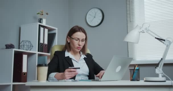 Mujer de negocios de compras en línea con el ordenador portátil en la oficina. Jovencita sostiene tarjeta de crédito bancaria en la mano y compra desde el ordenador . — Vídeos de Stock