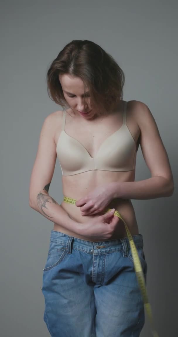 Женщина, стоящая перед зеркалом в джинсах, измеряет талию после потери веса. Концепция потери веса и диеты. Вертикальный шот — стоковое видео