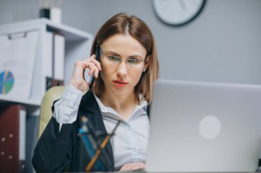 Genç bir kadın bilgisayarını kullanarak masada oturuyor, telefona cevap veriyor, telefonla konuşuyor, gülümsüyor cep telefonuyla konuşuyor, ofiste bilgisayarla çalışırken cep telefonunda hoş sohbetler ediyor.