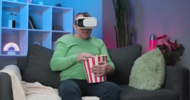 Senior Caucasian Man watching a video using a virtual reality headset Sitting on Sofa eating popcorn in a room