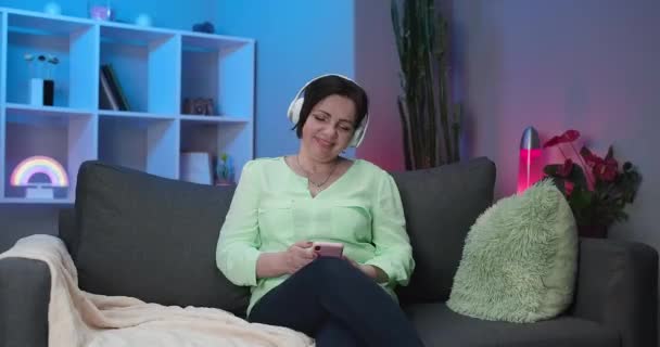 Happy Senior Woman w słuchawkach Słuchanie muzyki w domu. Starsza kobieta nosząca bezprzewodowe słuchawki Słuchając ulubionej piosenki i uśmiechu Happy. Technologia, ludzie starzy i koncepcja stylu życia. — Wideo stockowe