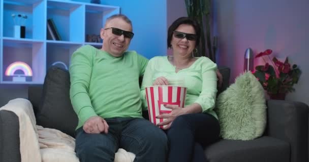 Пожилые мужчины и женщины смотрят телевизор вместе, сидя дома на диване и смотря увлекательный фильм в 3D очках. Пара пожилых людей в 3D очках и едят попкорн . — стоковое видео
