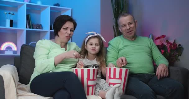 Starší kavkazský pár sedí doma na gauči s pětiletou vnučkou, usmívá se, dívá se na televizi a jí popcorn. — Stock video