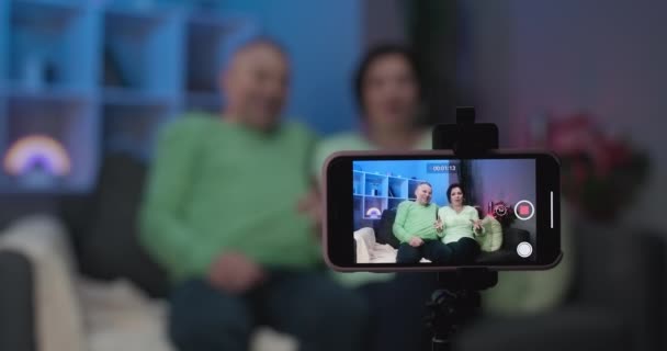 Casal de idosos usando videoconferência smartphone com neto enquanto deitado no sofá na sala de estar em casa. Desfrutando de tempo estilo de vida família sênior. Retrato olhando para a câmera — Vídeo de Stock