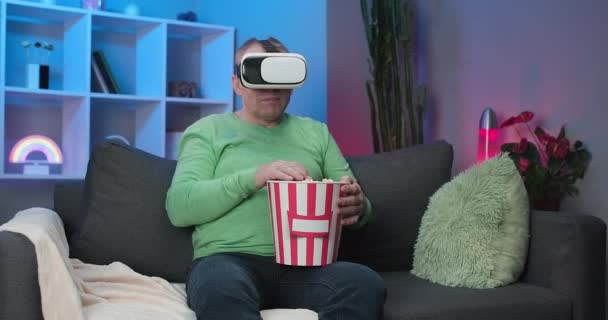 Älterer Mann mit Virtual-Reality-Brille isst Popcorn, während er zu Hause auf dem Sofa sitzt und Film schaut. — Stockvideo