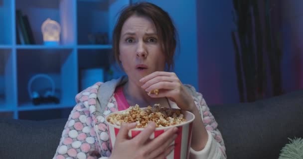 Портрет американской женщины с попкорном, сидящей на диване и смотрящей что-то страшное во время еды попкорна и страха — стоковое видео