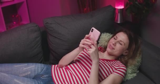Relaxed girl holding smart phone using mobile apps watching funny video laughing lying on couch, smiling lazy young woman having fun chatting in social media resting on sofa at home — Wideo stockowe