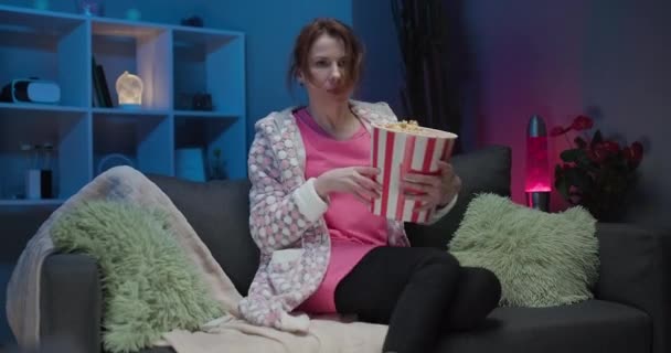 Mujer sentada en el sofá de la sala de estar con palomitas de maíz y canales cambiantes con mando a distancia mientras ve la televisión — Vídeos de Stock