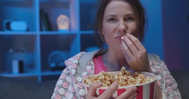 Cheerful woman sitting on the sofa in the living room with popcorn and watching TV — Stock video