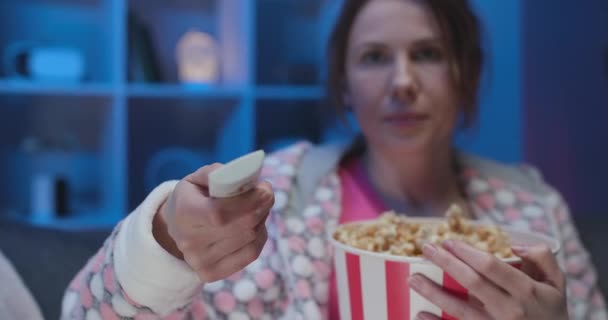 Mooie vrolijke vrouw zittend op de bank in de woonkamer met popcorn en wisselkanalen met een afstandsbediening terwijl je tv kijkt — Stockvideo