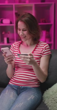 Akşamları kanepede oturan güzel beyaz bir kadın internetten alışveriş yaparken akıllı telefonu dinliyor ve kredi kartıyla ödüyor.