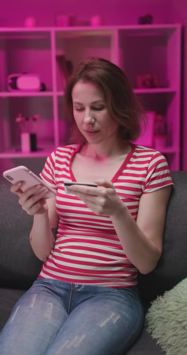 Vertical Shot Mujer joven haciendo compras en línea en el teléfono inteligente con tarjeta de crédito mientras está sentado en el sofá en la sala de estar . — Vídeo de stock