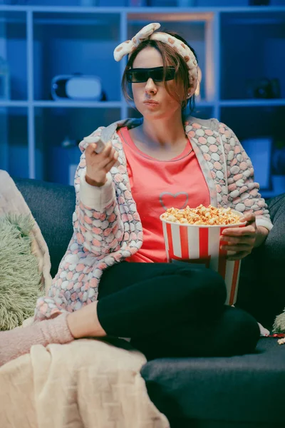 Woman on a soft couch eating popcorn, looking at the TV screen in 3D glasses. Woman eating popcorn in 3d glasses. — Stock Photo, Image