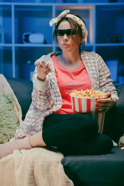 Женщина на мягком диване ест попкорн, смотрит на экран телевизора в 3D очках. Женщина ест попкорн в 3D-очках
.