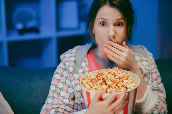 Woman with popcorn sitting on the sofa watching something scary while eating popcorn and being afraid