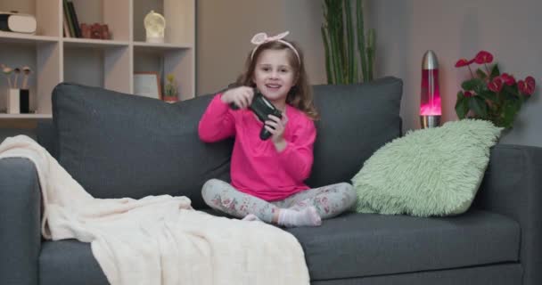 Pequeña niña sentada en el sofá jugando un video en la sala de estar en casa. Emocionado jugador mano chica sosteniendo joystick juego de consola usando un controlador inalámbrico. Divertirse, disfrutar de la expresión feliz — Vídeo de stock