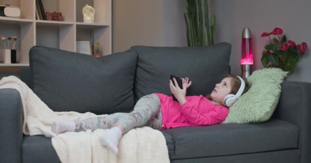 Glad liten flicka i trådlösa hörlurar kylande skrattar på soffan lyssna på favorit musik håller telefon med hjälp av mobilen online-spelare app njuta av lugn stämning bär hörlurar hemma — Stockvideo