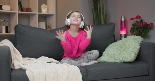 En liten flicka dansar på soffan medan hon lyssnar på musik i hörlurar hemma. Flicka klädd i hörlurar dansar, sjunger och flyttar till rytmen. — Stockvideo