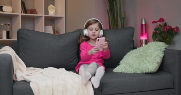 Cute little Caucasian girl sitting on sofa in home, smiling and listening to music in headphones on modern smartphone — Stock Video