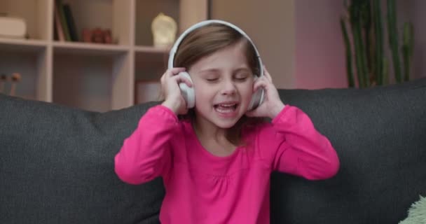 Cute Happy Little Girl Écoute de la musique sur des écouteurs sans fil. Petite fille drôle dansant, chantant et se déplaçant au rythme. Enfant portant des écouteurs . — Video