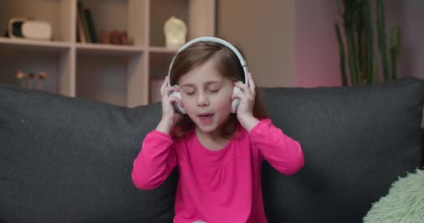 Meisje luistert naar muziek op draadloze koptelefoon. Grappig klein meisje dansen, zingen en bewegen om ritme. Kind draagt een koptelefoon. — Stockvideo