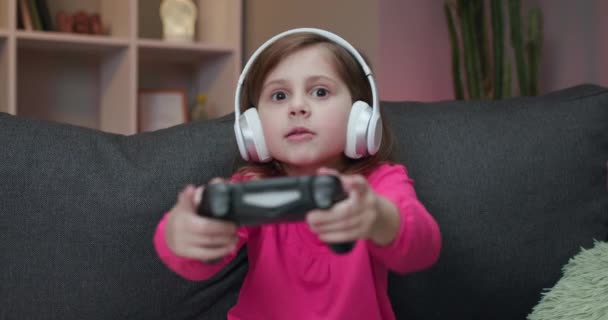 小女孩在家里玩电子游戏。兴奋的游戏玩家小女孩坐在沙发上，在游戏机上玩电子游戏，玩迷了路，用无线控制器 — 图库视频影像