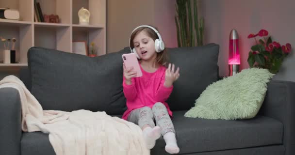 ヘッドフォンの女の子はソファで踊り、スマートフォンのタッチスクリーンを使用しています。現代技術と幸せな子供時代のコンセプト — ストック動画
