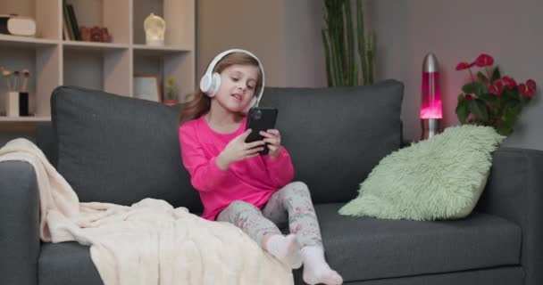 Liten flicka i bluetooth hörlurar njuter av musik med mp3-spelare på smartphone, sjunga med till sin favorit låt, svajande till takten samtidigt som du kopplar av på soffan — Stockvideo