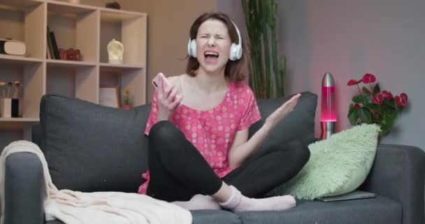 Радісна дівчина в навушниках танцює на дивані і використовує сенсорний екран смартфона. Сучасна концепція технології — стокове відео