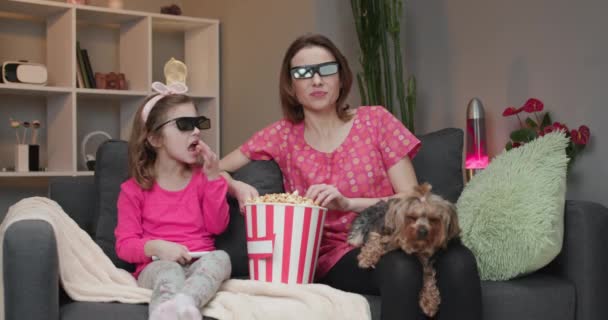 Mulher com uma menina e um cachorro vestindo óculos 3d assistindo TV e comendo pipocas. Tempo em família relaxar com criança menina no sofá no conceito de sala de estar — Vídeo de Stock