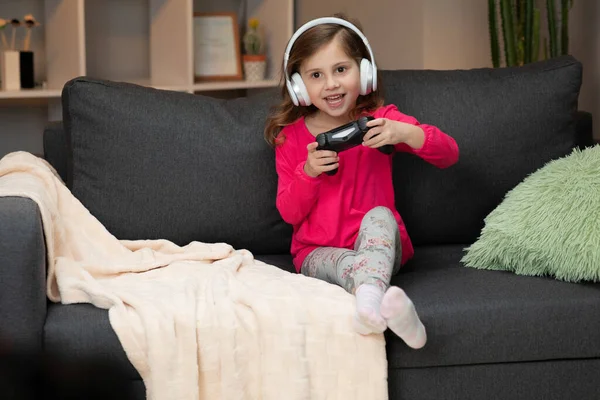 Jonge kleine meid zit op de bank en speelt een video in de woonkamer thuis. Opgewonden gamer meisje hand houden joystick spelen console spel met behulp van een draadloze controller. Plezier hebben, genieten van vrolijke expressie — Stockfoto