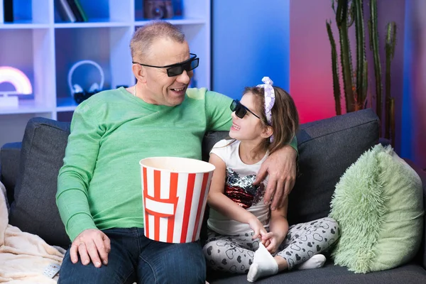 Oudere man met een klein meisje die 3D-bril draagt, tv kijkt en popcorn eet. Senior, oude generatie, grandphather familietijd ontspannen met jong meisje kind op de bank in woonkamer concept — Stockfoto