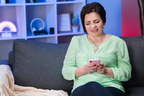 Senior Caucasian Woman with Short Black Hair Sitting on Couch at Home, Holding Invisible Smartphone in Hand and Chatting with Someone