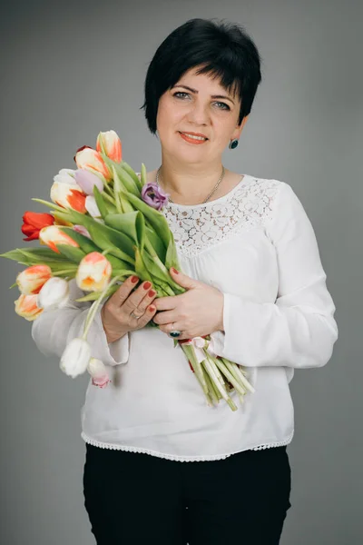 Attractive woman holds bouquet of flowers tulips in her hands gift soft toy looking at camera and smiling. Woman with tulip bouquet. Woman with gifts.