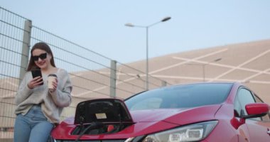 Elektrikli araba şarjı. Kadın elektrikli arabasının yanında duruyor ve aracın şarj olmasını bekliyor..