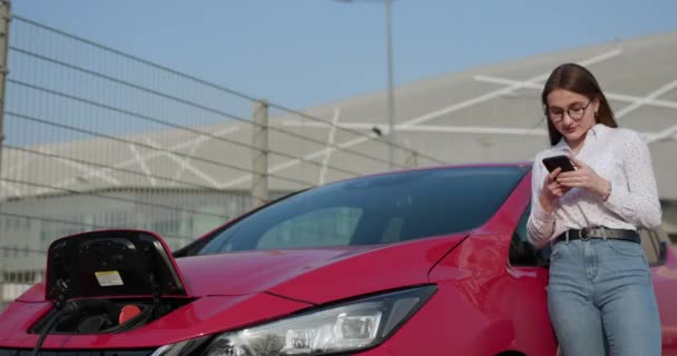 Ragazza sta con il telefono vicino alla sua auto elettrica rossa e aspetta quando il veicolo verrà caricato — Video Stock