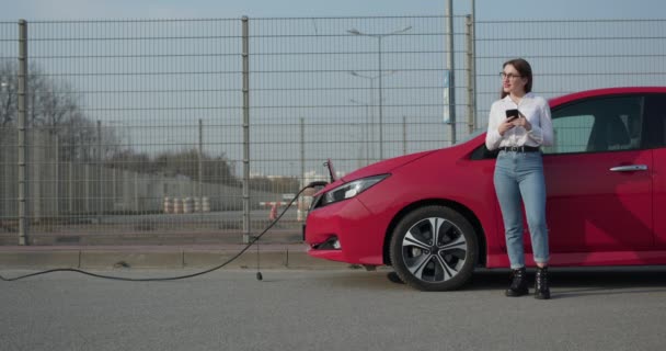 Girl stands with phone near her red electric car and waits when vehicle will charged. Ecological car connected and charging batteries. Plugging in power cord to an electric car. — Stock Video