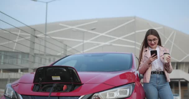 A menina usa a bebida do café ao usar o telefone esperto e a fonte de alimentação de espera conecta-se aos veículos elétricos para carregar a bateria no carro — Vídeo de Stock