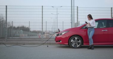 Ekolojik Araba Bağlı ve Şarj Bataryaları. Kız Akıllı Telefon Kullanırken Kahve İçiyor ve Elektrikli Araçlara Bağlanarak Araçtaki Bataryayı Şarj Etmek İçin Bekliyor.