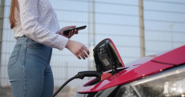 O processo de recarga elétrica de carros chega ao fim. Menina caucasiana usando telefone inteligente e esperando fonte de alimentação conectar a veículos elétricos — Vídeo de Stock