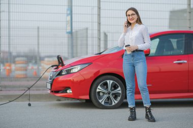 Elektrikli araba caddede şarj oluyor. Ekolojik Araba Bağlı ve Şarj Bataryaları. Akıllı Telefon ve Bekleme Güç Aracını Şarj Etmek İçin Elektrikli Araçlara Bağlanırken Kahve İçeceği Kullan