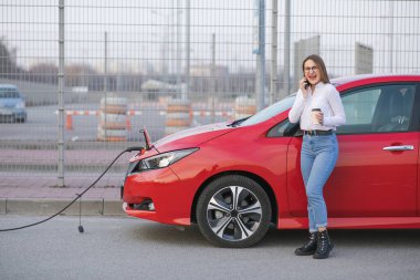 Ekolojik Araba Bağlı ve Şarj Bataryaları. Kız Akıllı Telefon Kullanırken Kahve İçiyor ve Elektrikli Araçlara Bağlanarak Araçtaki Bataryayı Şarj Etmek İçin Bekliyor.