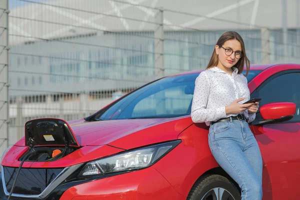 Dívka používající chytrý telefon a čekající napájení připojit k elektrickým vozidlům pro nabíjení baterie v autě. Ekologické auto připojeno a nabíjení baterií — Stock fotografie